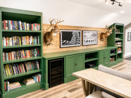 Custom green built-in cabinetry featuring floor-to-ceiling bookshelves, lower storage cabinets, a wood accent wall, integrated entertainment storage, and display shelving in a finished bonus room.