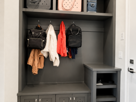 Custom gray mudroom built-in featuring upper storage cubbies, coat hooks, bench seating, ventilated storage cabinets, and open shoe shelving near an entryway.