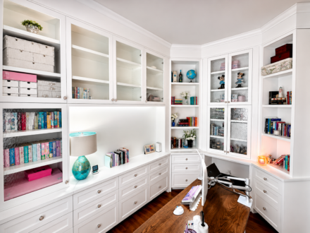 Custom white built-in home office featuring floor-to-ceiling cabinetry, glass display doors, extensive drawer storage, open shelving, and a rich wood desk in a bright, elegant workspace.