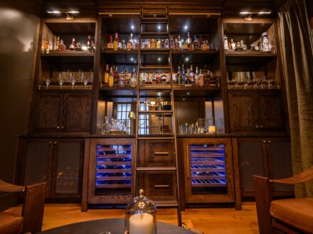 Custom white oak bourbon bar featuring floor-to-ceiling cabinetry, a rolling library ladder, integrated wine refrigerators, brass gallery rails, display lighting, mesh cabinet doors, and premium liquor storage.